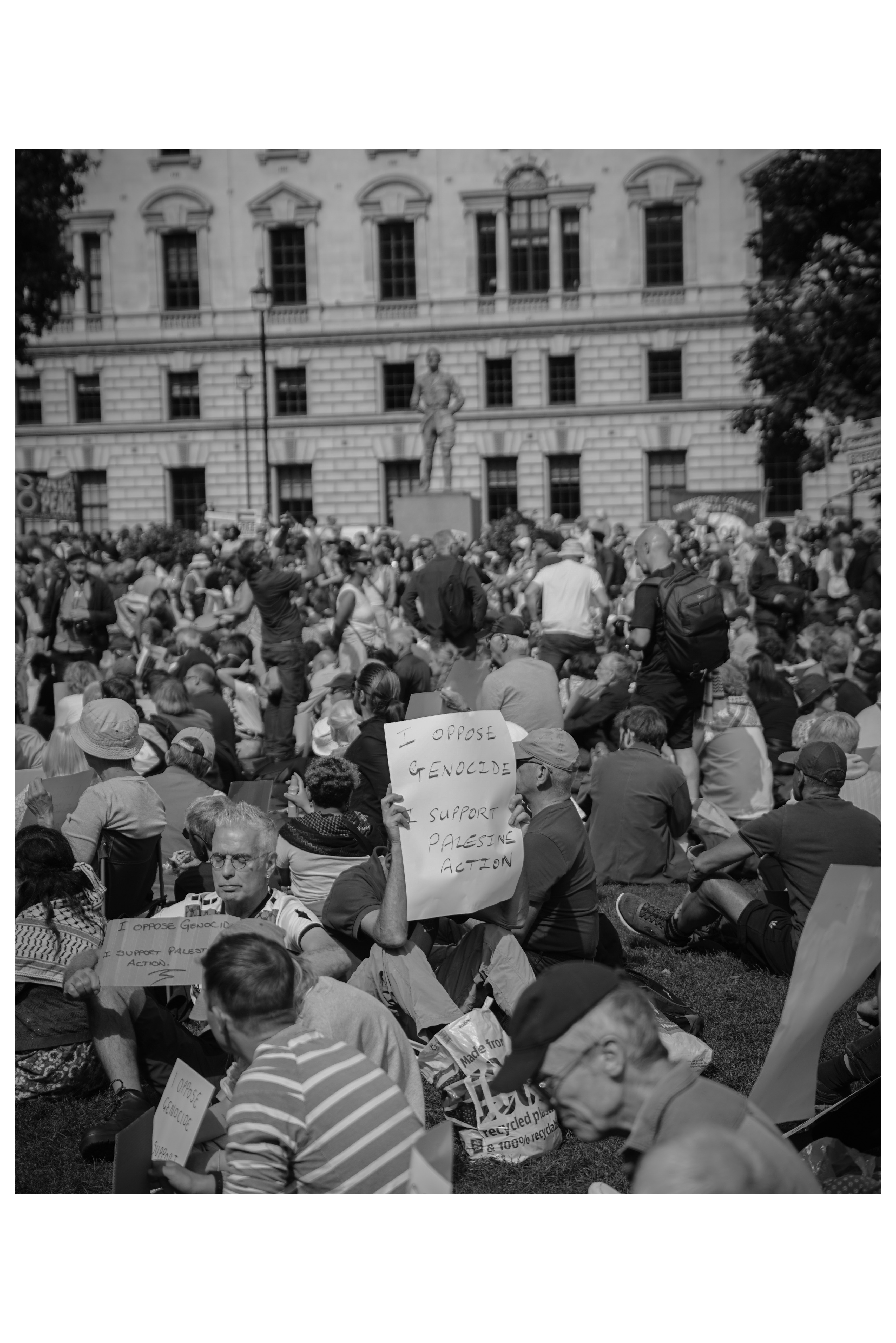 A protester holds a handwritten sign reading 'I Oppose Genocide, I Support Palestine Action' among a large crowd of protesters gathered in Parliament Square, London.